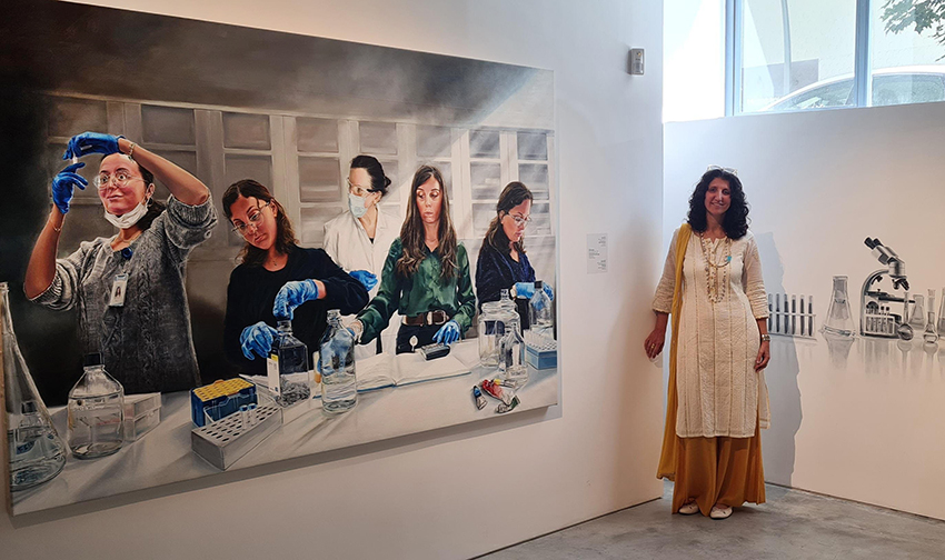 שירלי סיגל לצד תמונת המדעניות בתערוכה. המוזיאון הרחיב את התמונה גם לקיר הסמוך באמצעות ציור של כלי עבודה אופייניים למחקרים בביולוגיה מולקולרית | צילום: באדיבות שירלי סיגל