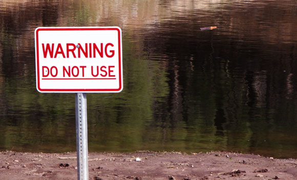הזיהום במים משמיד אוכלוסיות דגים ודו-חיים, ומסכן את מקורות מי השתייה. אגם עם שלט אזהרה. צילום: Shutterstock